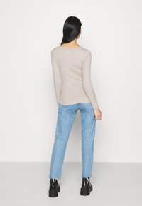 Pitkä mustahiuksinen nainen, joka pitää päällään beigeä ribbineulosta villapaitaa, vaaleansinisiä repaleisia farkkuja ja mustia korollisia saappaita, seisoo valkoista taustaa vasten.