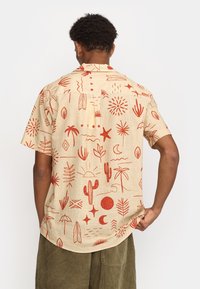 Kortärmige beige shirt met rode grafische patronen van sterren, zon, cactussen, palmbomen en golven. Met kraag, relaxte pasvorm.