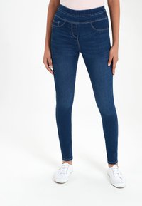 Jeggings bleu foncé à taille haute, présentant deux poches avant, coupe slim et détails de couture subtils. Portés avec des baskets blanches.