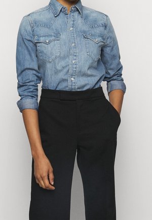 Osoba ubrana w dopasowaną, zapinaną na guziki koszulę denimową z podwiniętymi rękawami i czarne spodnie z wysokim stanem, jedna ręka w kieszeni, na jednolitym tle.