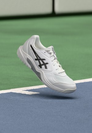 Biela športová obuv ASICS s čiernym logom vznášajúca sa nad modro-zeleným povrchom tenisového dvorca pri základnej čiare.