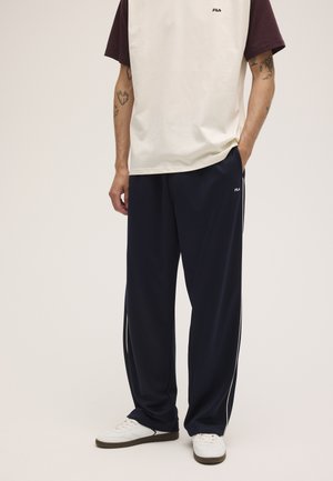 Hombre con camiseta blanca y granate, pantalones deportivos FILA azul marino y zapatillas blancas, de pie con las manos en los bolsillos.
