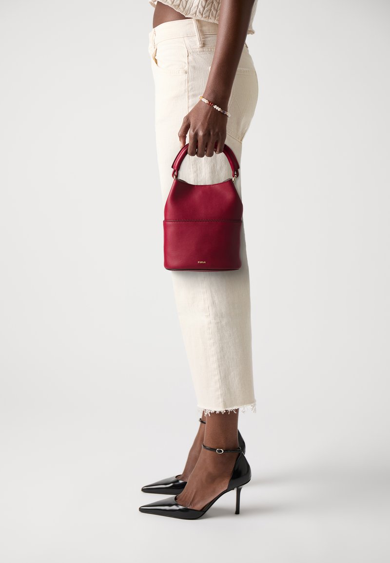 Sac à main en cuir rouge avec un dessus incurvé et une courte poignée. Présente une texture lisse et des coutures discrètes. Associé à un jean blanc court et des talons noirs.