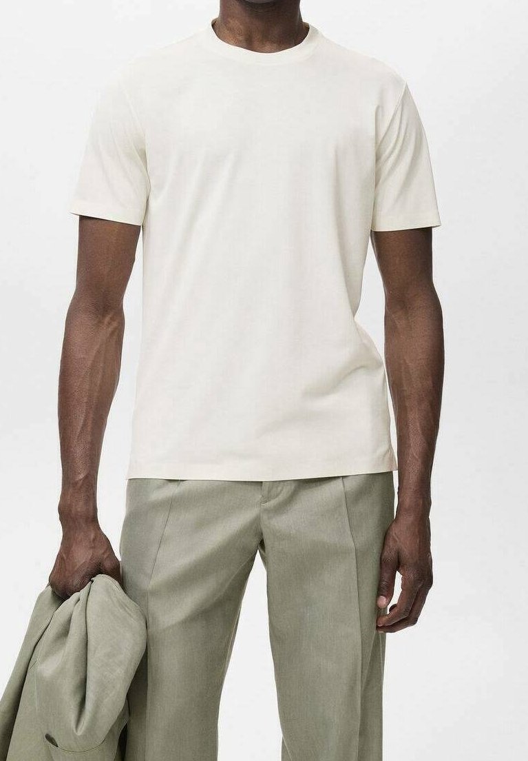 Homme portant un t-shirt blanc uni à manches courtes et un pantalon gris clair, tenant une veste assortie gris clair dans sa main gauche devant un fond uni.