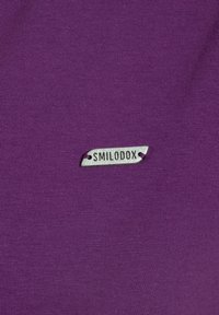 Tela de algodón púrpura con una textura suave, que presenta una pequeña etiqueta metálica que dice "SMILODOX."