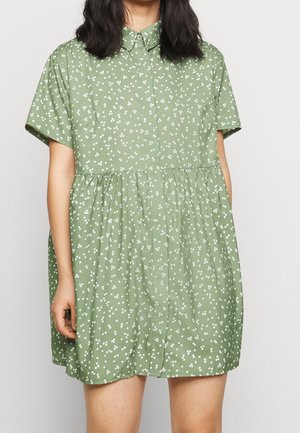 Femme portant une robe vert clair à manches courtes avec des petits pois floraux blancs et un col, debout devant un fond uni.