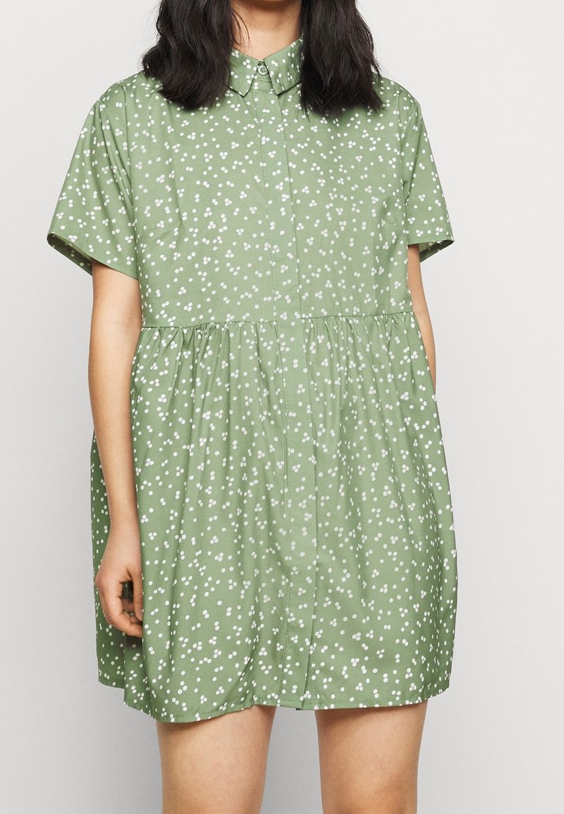Femme portant une robe vert clair à manches courtes avec des petits pois floraux blancs et un col, debout devant un fond uni.