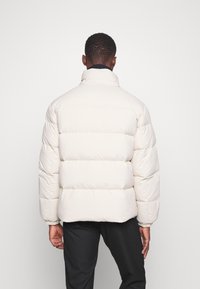 Hombre de espaldas con una chaqueta acolchada beige clara y pantalones negros, contra un fondo blanco liso.