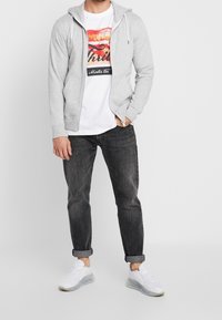 Lichtgrijze hoodie met rits over een wit T-shirt met een kleurrijke lippenprint, gecombineerd met opgerolde donkere jeans en witte sneakers.