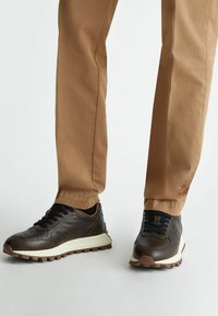 Sneakers in pelle marrone con lacci neri, suola intermedia bianca e suola esterna in gomma marrone texturizzata. Indossati con pantaloni beige.