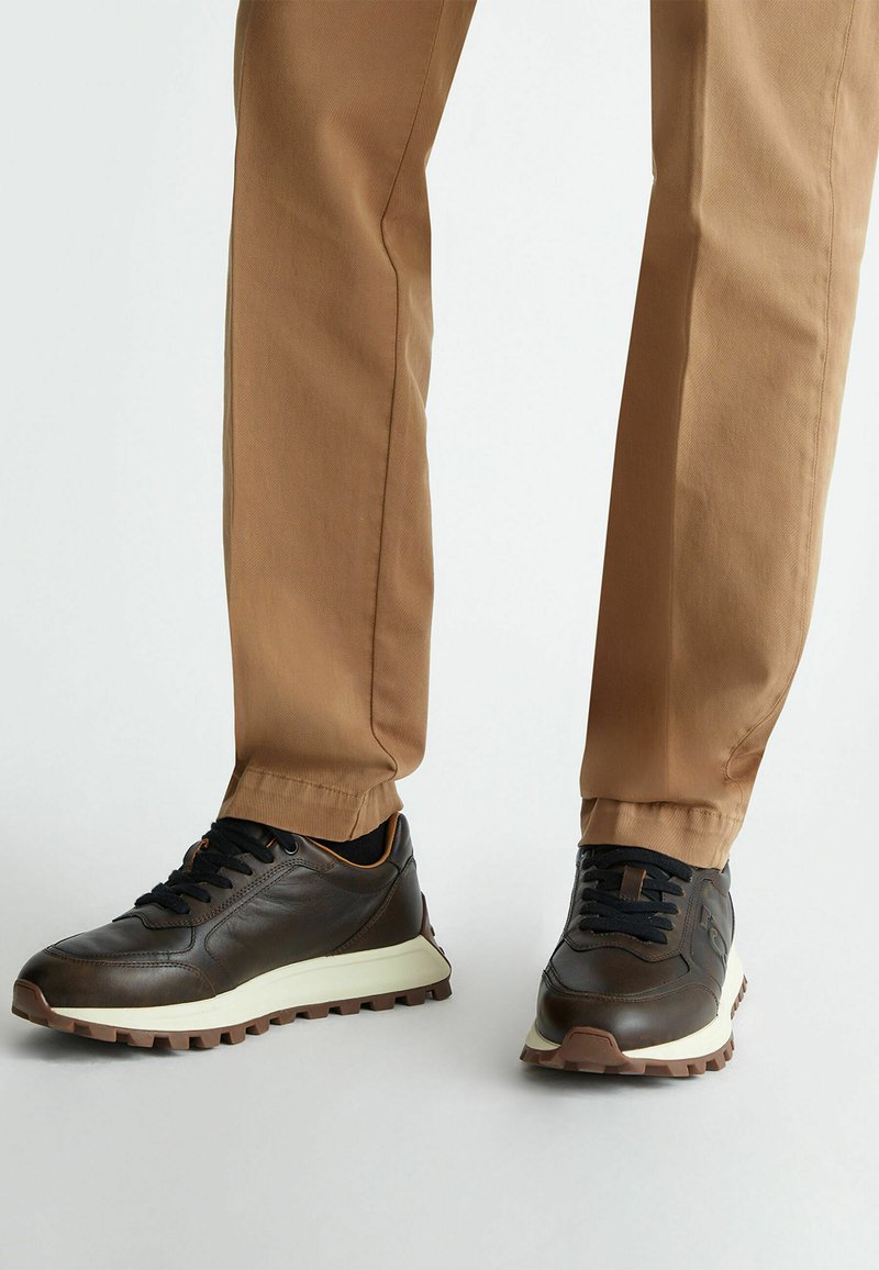 Sneakers in pelle marrone con lacci neri, suola intermedia bianca e suola esterna in gomma marrone texturizzata. Indossati con pantaloni beige.