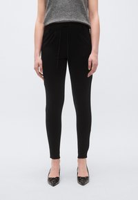 Pantaloni negri ajustați, cu talie cu șnur, buzunare laterale și textură fină, asortați cu pantofi negri cu vârf ascuțit.