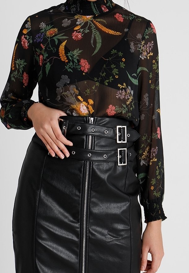 Blouse noire fleurie transparente à manches longues rentrée dans une jupe en cuir noire taille haute avec double boucle et fermeture éclair devant.