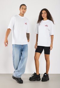 Hvide oversized t-shirts med et lille farvet logo, parret med lyseblå baggy jeans og sorte sneakers. Kvinden har sorte cykelshorts på.