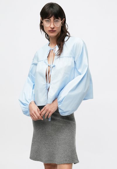 Ανοιχτό γαλάζιο κοντό τοπ με φουσκωτά μανίκια, με κλείσιμο μπροστά με δέσιμο και ανοιχτό λαιμόκοψη. Φορεμένο πάνω από μια γκρι φουσκωτή φούστα.