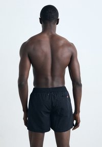 Black swim shorts with an elastic waistband, a rear pocket featuring a small red logo, and a lightweight, smooth fabric texture.