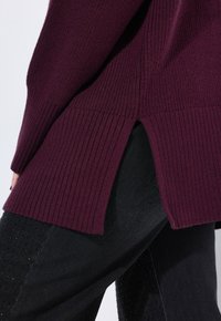 Burgundowy sweter z dzianiny o prążkowanej fakturze, z bocznymi rozcięciami. Połączony z czarnymi spodniami z perforowanymi detalami.