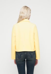 Κοντό σακάκι κίτρινου μείγματος μαλλιού με γιακά με εγκοπή και μακριά μανίκια, συνδυασμένο με σκούρο τζιν τζιν παντελόνι με διακοσμητικές πίσω τσέπες.