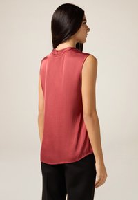 Ärmelloses Oberteil in einem satten Burgunderrot, aus glattem, glänzendem Material mit hohem Hals und abgerundetem Saum, kombiniert mit schwarzen Hosen.