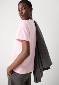 Short-sleeve pink cotton t-shirt paired with gray textured fabric draped over the shoulder. Model poses with hand in pocket.