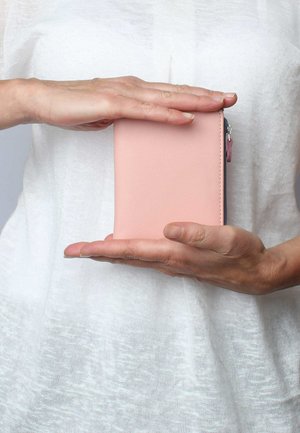 Rosa Lederbörse, rechteckige Form, glatte Textur, zwischen Händen gehalten, mit einem Reißverschluss, der von einem kleinen farbigen Akzent gekrönt wird.