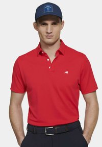 Rode polo shirt van een gestructureerde stof, voorzien van een kraag en een drieknopen-sluiting. Gecombineerd met een marineblauwe pet met een logo-patch.