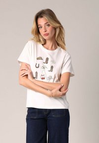 T-shirt en coton blanc avec un imprimé graphique de diverses bottes, chapeaux, un fer à cheval et des cerises. Manches courtes, coupe décontractée.
