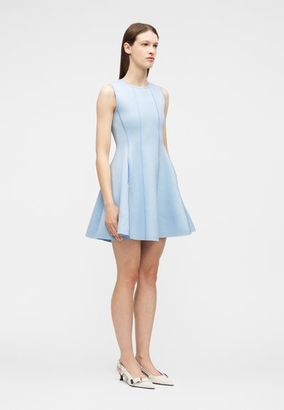 Femme debout portant une robe évasée sans manches bleu clair et des escarpins blancs à bouts pointus et bride arrière, devant un fond uni.