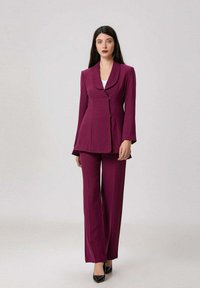 Femme portant un tailleur prune ajusté avec une veste à col châle, associée à des escarpins noirs à bout pointu, se tenant devant un fond uni.