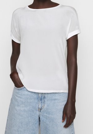 Personne à la peau foncée portant un haut blanc uni à manches courtes et un jean en denim bleu clair taille haute, une main dans la poche.