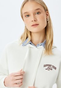 Sudadera crema con un bolsillo frontal y cordón ajustable. Presenta texto marrón que dice "SOPORTA LO LOCAL". Lleva puesta sobre una camisa a rayas azul y blanca.