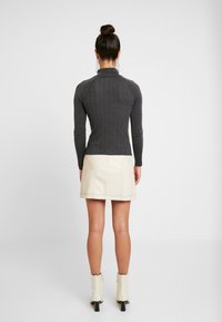 Femme avec les cheveux en chignon portant un pull ajusté gris foncé à côtes, une mini-jupe en cuir crème, des bottines blanches, debout dos à la caméra sur fond blanc.