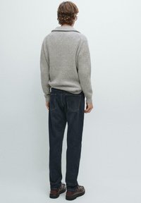 Maglione grigio a maglia con texture a coste, abbinato a jeans di denim blu scuro. Il modello indossa scarpe marroni, visto di spalle.