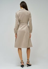 Vestido beige hasta la rodilla con cuello, mangas largas y cintura ceñida con un cinturón. Presenta una tela suave y acentos de botones dorados en los puños.