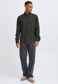 Ung mand stående, iført en grøn- og sortternet skjorte med knapper, løse sorte jeans og brune sneakers, kiggende til venstre.