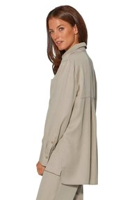 Beige Langarmhemd mit Kragen, ausgestattet mit einer Tasche, Knopfdetails an den Manschetten und einem lässigen Schnitt. Sanfte Textur, leicht übergroßes Design.