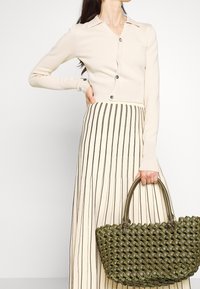 Bolso de mano verde tejido con forma redondeada y asas dobles, combinado con un cárdigan beige claro y una falda midi a rayas, que cuenta con un cierre de botones.