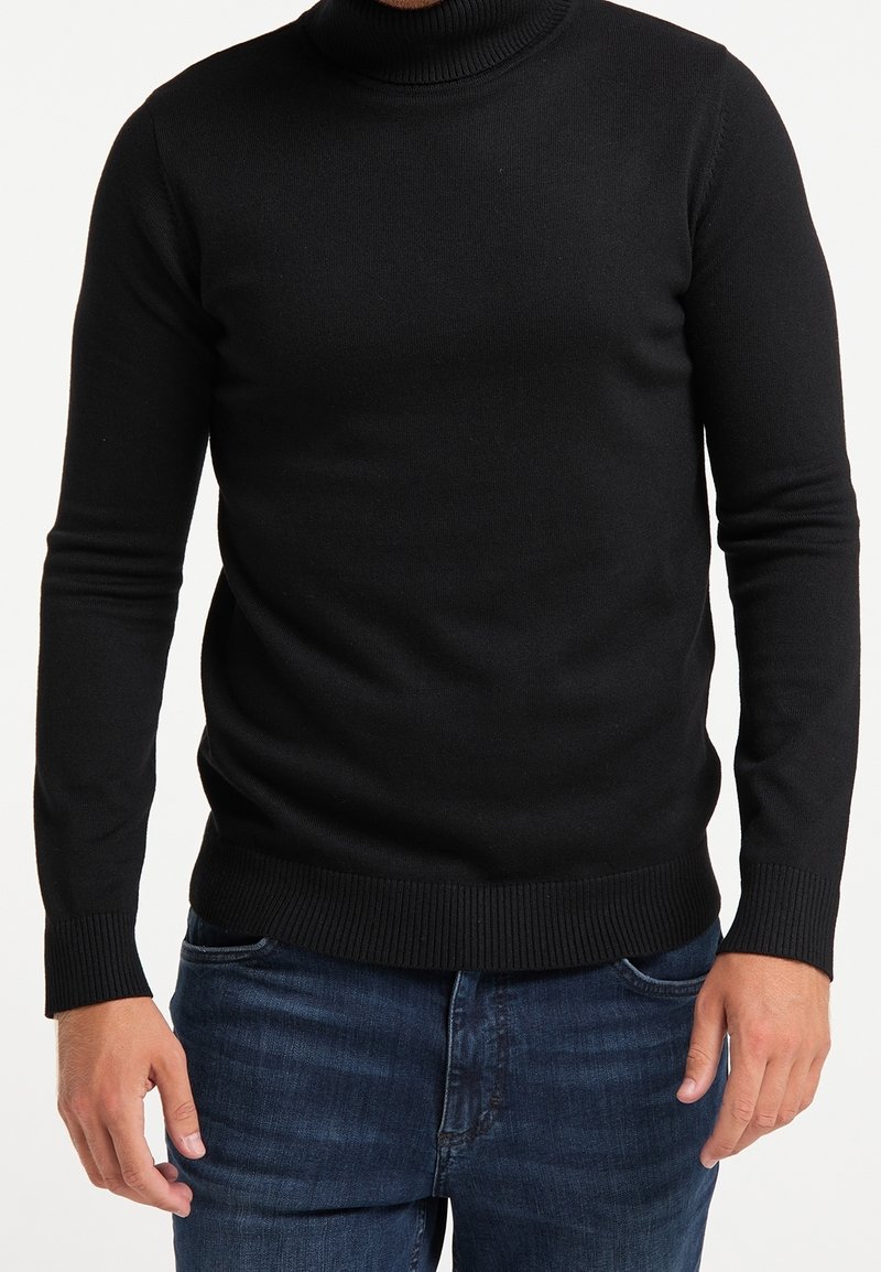 Homme portant un pull noir à col roulé ajusté et un jean bleu, debout devant un fond blanc uni.