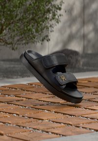 Μαύρο σανδάλι τύπου σλάιντ κατασκευασμένο από λείο δέρμα, με δύο ρυθμιζόμενους ιμάντες με velcro και διακοσμητικό λογότυπο σε χρυσή απόχρωση. Διαθέτει μαλακή, επενδεδυμένη σόλα για άνεση.