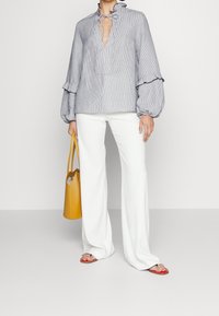 Blouse légère rayée avec un col en V et un détail noué, dotée de manches volumineuses et de poignets à volants. Associée à un pantalon blanc évasé et un sac jaune.