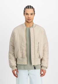 Beige bomber jacket με βαμβακερή υφή, φερμουάρ, ελαστικούς καρπούς και δύο πλαϊνές τσέπες, φορεμένο πάνω από ένα πράσινο πουκάμισο και ανοιχτόχρωμα τζιν.