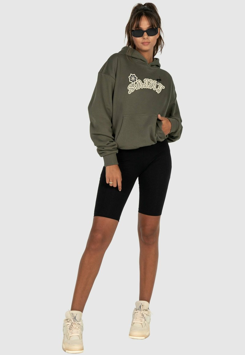 Oliven grøn hætte sweatshirt med "SAINT" logo, parret med sorte cykelshorts og hvide højtaljede sneakers. Glat tekstur, afslappet pasform.