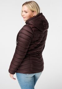Bruine pufferjack met een capuchon, voorzien van een doorgestikt ontwerp en ribbelkraag. Draagt over lichtblauwe jeans, wat een getailleerd silhouet laat zien.