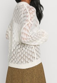 Bílý pletený svetr s texturovaným, otevřeným vzorem a dlouhými rukávy, doplněný hnědou skládanou sukní, která předvádí lehkou a vzdušnou látku.