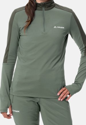 Woman wearing olive green long-sleeve athletic top and pants with thumb holes and Vaude logos, standing with one hand on hip.