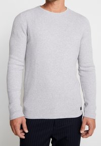 Ljusgrå, ribbad stickad långärmad tröja med rund halsringning och figurnära form, som har en liten logotag i kanten.