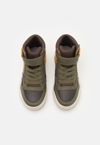 Un par de zapatillas altas en verde y marrón con suelas blancas y acentos naranjas, vistas desde arriba sobre un fondo blanco.