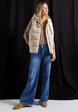 Beige zachte vest met een capuchon, gelaagd over een lichtbruine top, gecombineerd met wijde blauwe jeans en tan instapschoenen, tegen een zwarte achtergrond.
