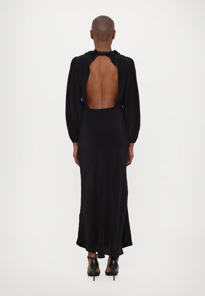 Juoda suknelė ilgomis rankovėmis, su iškirpte nugaroje, laisvo kirpimo ir tekstūruotu kaklu. Dėvima su juodais aukštakulniais bateliais.