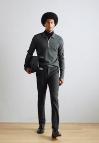 Temno siva zip zanka preko črnega puloverja s turtleneck ovratnikom, črne hlače in čevlji, drži črno torbo, stoji na lesenem tlaku ob svetli steni.
